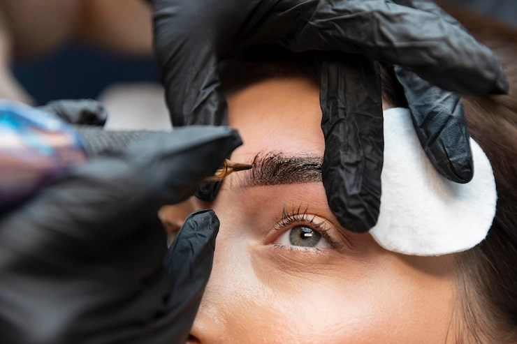 woman receiving eyebrow microblading treatment with gloved hands and tattoo machine
